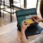 Seorang pengguna memegang kartu rekeningnya sambil bersiap melakukan transaksi online di perangkat digital, merepresentasikan proses pembayaran melalui bank transfer payment gateway, yang menghubungkan pembeli, merchant, dan bank untuk memastikan transaksi berlangsung aman dan efisien.